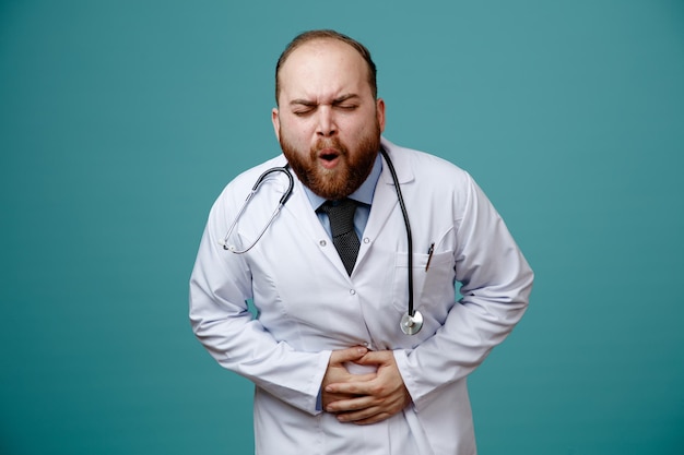 Aching young male doctor wearing medical coat and stethoscope around his neck keeping hands on belly with closed eyes isolated on blue background