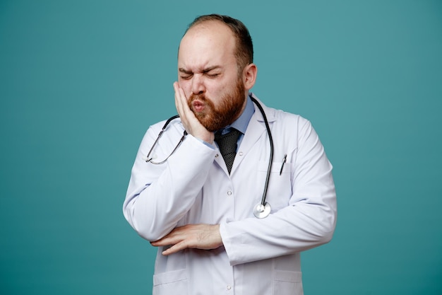 aching young male doctor wearing medical coat and stethoscope around his neck touching elbow keeping hand on cheek having toothache with closed eyes isolated on blue background