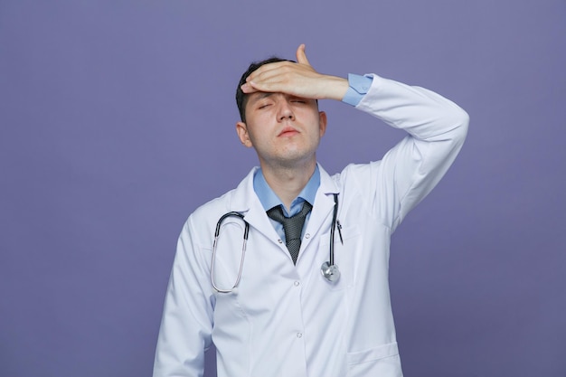 Aching young male doctor wearing medical robe and stethoscope around neck keeping hand on forehead with closed eyes having headache isolated on purple background