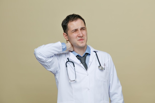 Aching young male doctor wearing medical robe and stethoscope around neck keeping hand behind neck with closed eyes isolated on olive green background