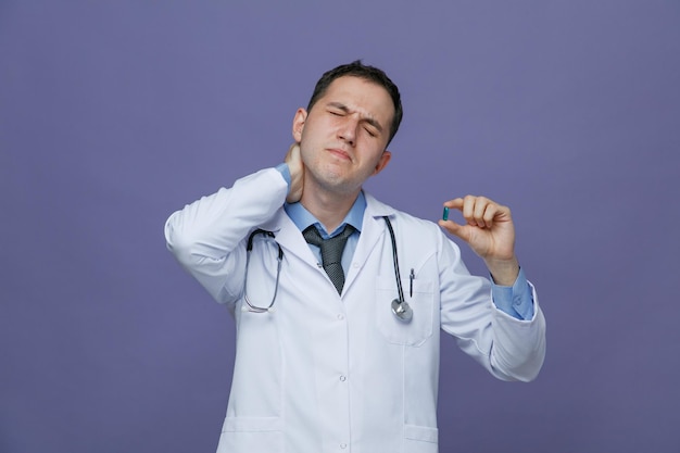 aching young male doctor wearing medical robe and stethoscope around neck showing capsule with closed eyes while keeping hand on neck isolated on purple background