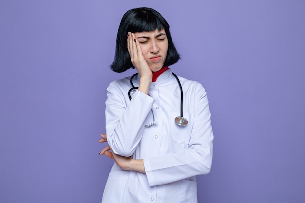 Aching young pretty caucasian woman in doctor uniform with stethoscope putting hand on face standing with closed eyes 