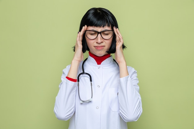 Dolorante giovane bella donna caucasica con gli occhiali che indossa l'uniforme del medico con lo stetoscopio che mette le mani sulla tempia in piedi con gli occhi chiusi