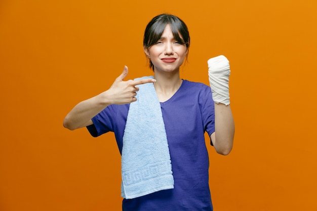Aching young sporty woman wearing tshirt showing her hand pointing at it looking at camera with towel on shoulder with wrist wrapped with bandage isolated on orange background