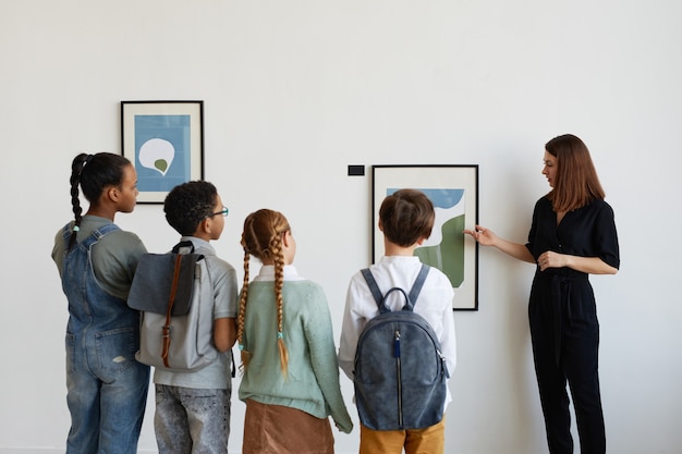 Achteraanzicht bij diverse groep kinderen die naar vrouwelijke gids luisteren tijdens een bezoek aan een moderne kunstgalerie, kopieer ruimte