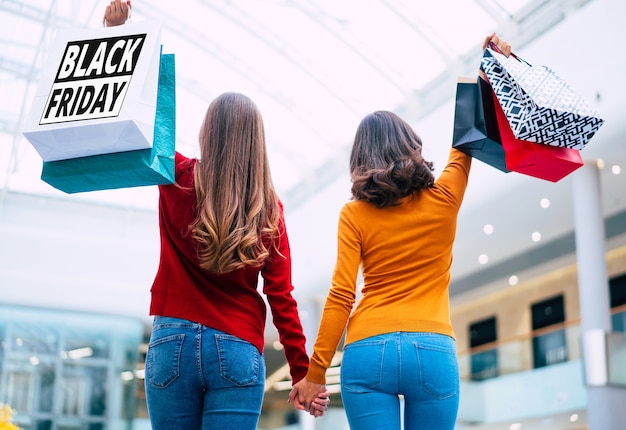 Achteraanzicht foto van twee jonge vrouwen in kleurrijke kleding met boodschappentassen in handen met print erop van zwarte vrijdag.