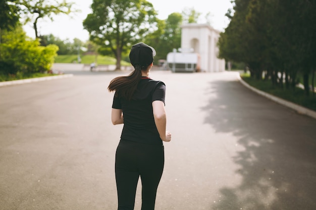 Achteraanzicht jonge atletische brunette meisje in zwart uniform en cap training, sportoefeningen doen en hardlopen, recht op pad in stadspark buitenshuis kijken