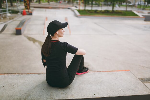 Achteraanzicht jonge atletische brunette vrouw in zwart uniform en pet met koptelefoon luisteren naar muziek rusten en zitten voor of na het hardlopen, training in stadspark buitenshuis