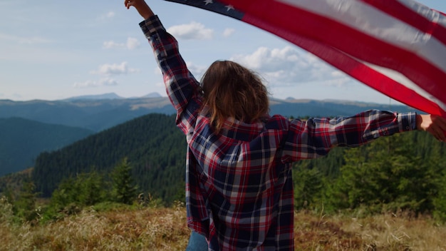 Foto achteraanzicht jonge vrouw met amerikaanse vlag in handen staande op de top van de berg vrouwelijke wandelaar zwaaiende nationale vlag van de vs buiten patriottisch meisje viert ons onafhankelijkheidsdag op vakantie