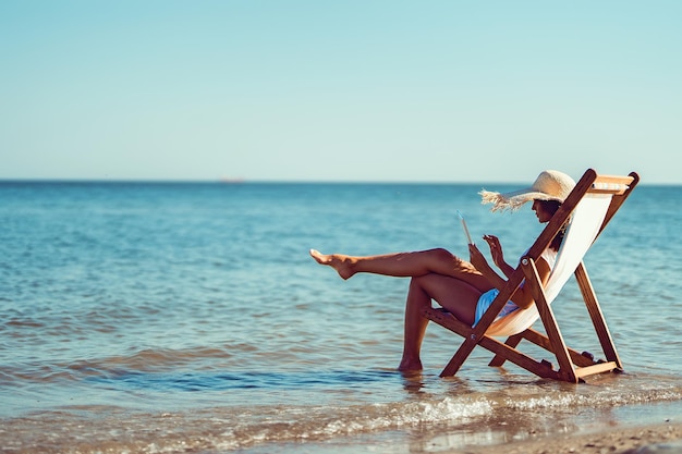 Achteraanzicht meisje in zonnehoed werken op laptop terwijl liggend op het strand chaise longue aan de kust