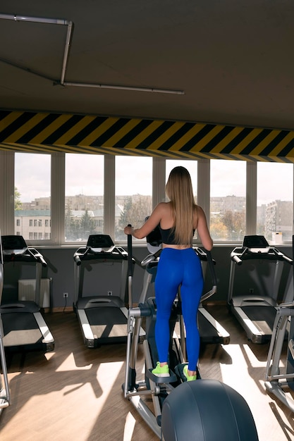 Achteraanzicht op mooi meisje met lang blond haar treinen in de sportschool. Elliptische trainer voor cardio.