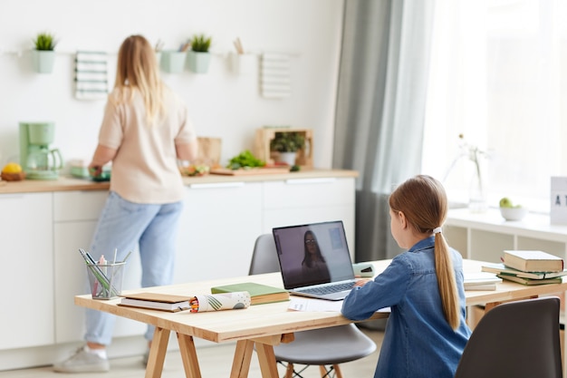 Achteraanzicht portret van meisje met laptop tijdens online les met tutor of leraar zittend aan een bureau in gezellige keuken interieur met moeder op achtergrond, kopie ruimte