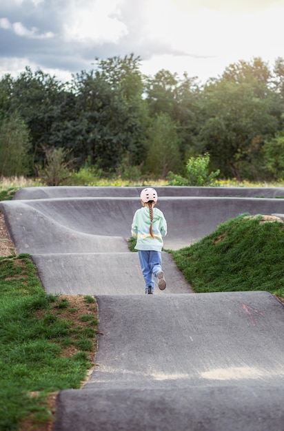 Achteraanzicht portret van schattig klein Kaukasisch schoolmeisje draag helm geniet van plezier scooter rijden op geasfalteerde trackxAin straatpark buitenshuis op zonnige dag Gezonde sportactiviteiten voor kinderen buiten