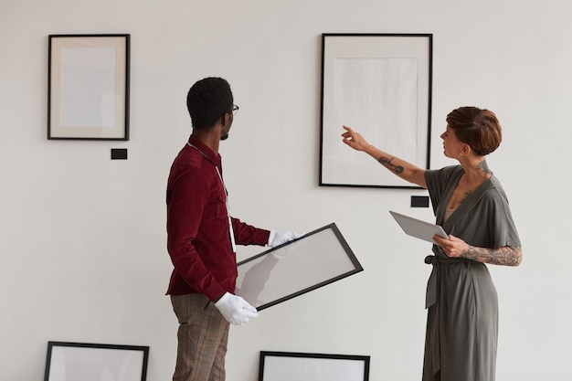 Achteraanzicht portret van vrouwelijke kunstgalerie manager instrueren werknemer schilderij frames op witte muur opknoping tijdens het plannen van tentoonstelling in museum,