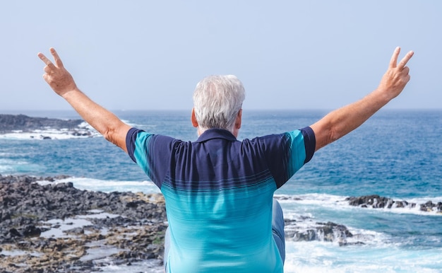 Achteraanzicht portret van witharige senior man met blauwe t-shirt genieten van buiten vakantie op zee Oudere man met uitgestrekte armen kijken naar horizon over water Vakantie of pensioen concept