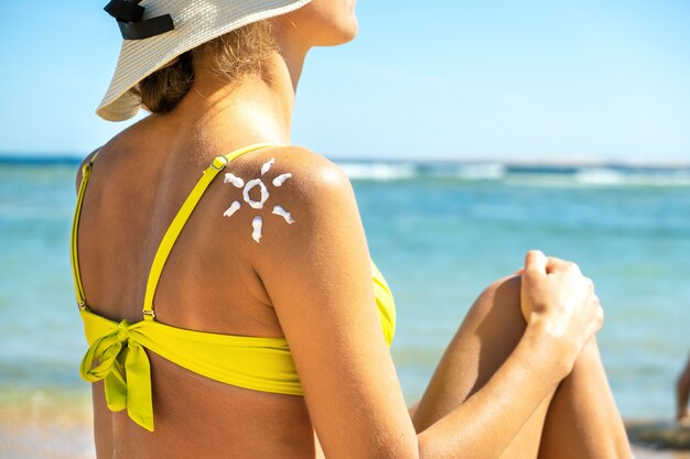 Achteraanzicht van de jonge vrouw die op het strand met zonnebrandcrème in de vorm van de zon op haar schouder looien.