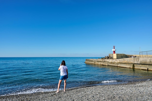 Achteraanzicht van de vrouw in T-shirt en korte broek op het strand kijkt op zee en rode vuurtoren op de pier tegen de blauwe lucht op zonnige dag