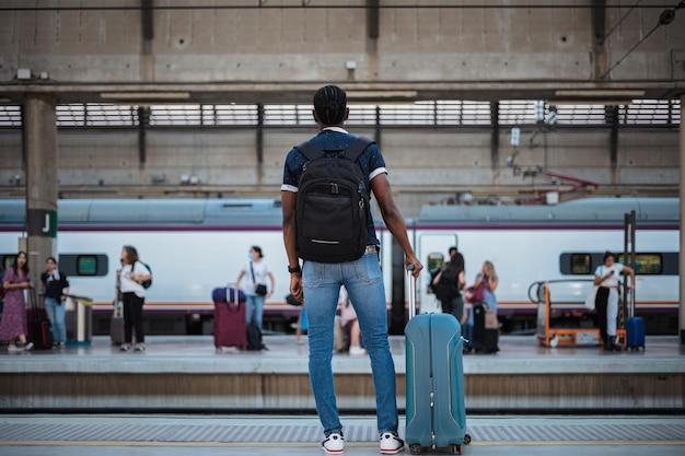 Achteraanzicht van een Afrikaanse man die op de trein wacht Hij houdt een blauwe koffer vast en heeft een zwarte tas