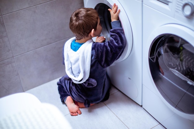 Achteraanzicht van een baby zit op de grond en kijkt in de wasmachine Het kind in zachte pyjama kijkt zorgvuldig naar het wassen