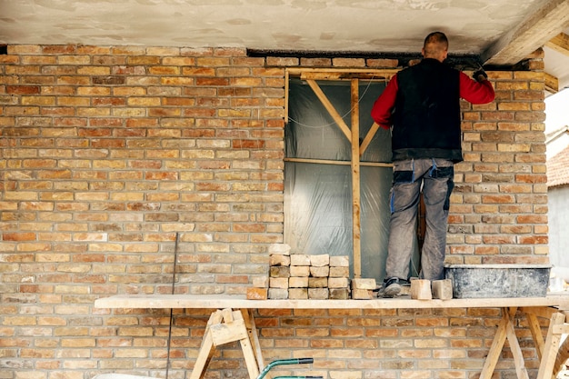 Achteraanzicht van een bouwer die bakstenen op een bakstenen muur stapelt terwijl hij op een steiger staat