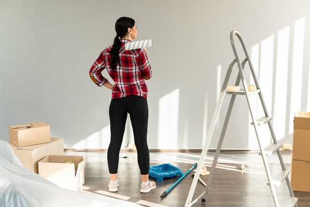 Achteraanzicht van een casual brunette vrouw die naar de muren kijkt terwijl ze haar woonkamer schildert in nieuwe appartementen