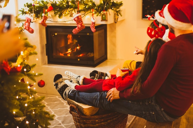 Achteraanzicht van een gelukkige familie die kerstkleding draagt en viert dat ze bij de open haard onder de versierde kerstboom zitten