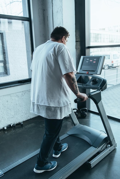 Achteraanzicht van een getatoeëerde man met overgewicht die op een loopband loopt in een sportcentrum