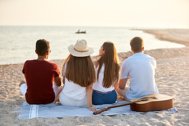 Achteraanzicht van een groep vrienden die samen op een handdoek op het witte zandstrand zitten tijdens hun vakantie en genieten van een zonsondergang boven de zee Mannen en vrouwen ontspannen aan de kust met gitaar Rustige scène