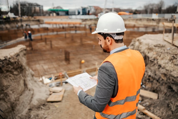 Achteraanzicht van een ingenieur die blauwdrukken op de bouwplaats bekijkt