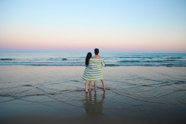 Achteraanzicht van een jong stel op het strand, samen gewikkeld in een handdoek, kijkend naar de zonsondergang