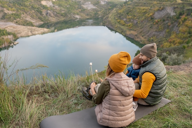 Achteraanzicht van een jonge hedendaagse backpackerfamilie die op een tapijt zit voor een prachtig meer omringd door bergen en marshmallows heeft