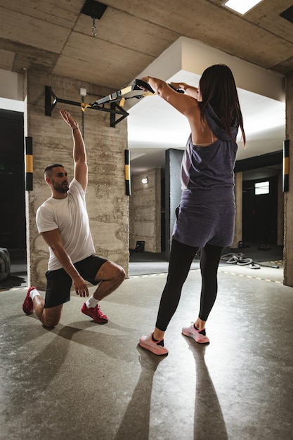 Achteraanzicht van een jonge knappe vrouw die aan het trainen is met een ophangriem in de sportschool, ondersteund door haar persoonlijke mannelijke trainer.