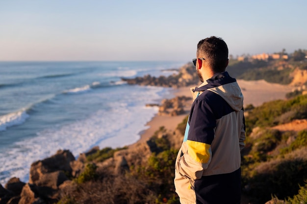 Achteraanzicht van een jonge man met jas en zonnebril kijkend naar de zee vanaf een klif naast het strand vrije tijd en ontspannen concept kopie ruimte voor tekst