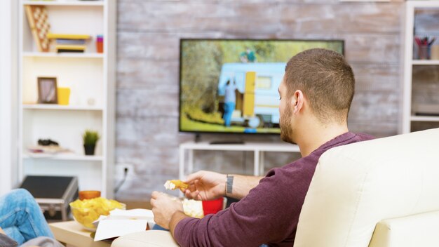 Achteraanzicht van een jonge man zittend op een comfortabele stoel die gebakken kip eet met zijn vriendin voor tv.