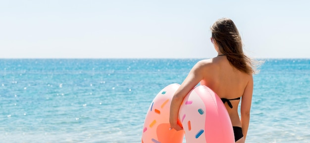 Achteraanzicht van een jonge vrouw die een rubberen ring van een donut vasthoudt en de zee in loopt Hete zomer