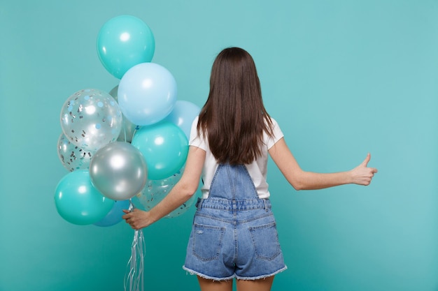 Achteraanzicht van een jonge vrouw in denim kleding met duim omhoog, vieren, met kleurrijke luchtballonnen geïsoleerd op blauwe turquoise muur achtergrond. vakantie verjaardagsfeestje, mensen emoties concept.