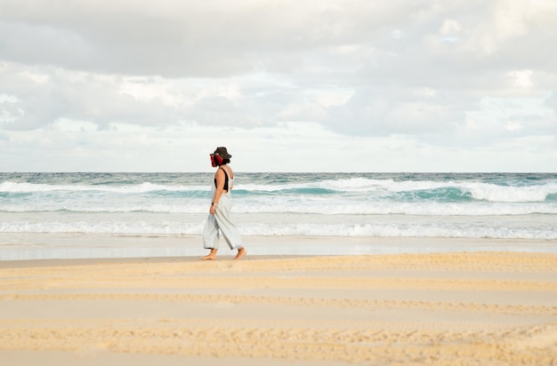 Achteraanzicht van een jonge vrouw met een hoed die alleen op een strand loopt en naar de oceaan kijkt Copy Space