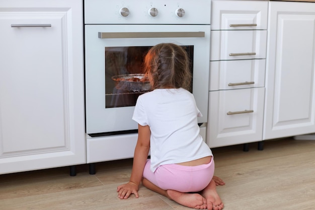 Achteraanzicht van een klein meisje dat in de buurt van de oven in de keuken zit en naar binnen kijkt door glas nieuwsgierig ongeduldig vrouwelijk kind dat wacht op koekjes die in het fornuis bakken en genieten van zelfgemaakt gebak