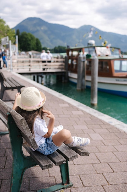 Achteraanzicht van een klein meisje met een strohoed, zittend op een bankje en kijkend naar de haven van een schip