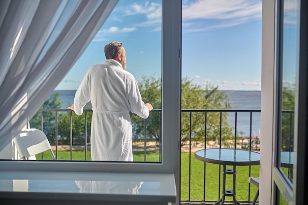 Achteraanzicht van een man in de witte badstof badjas leunend op de balustrade