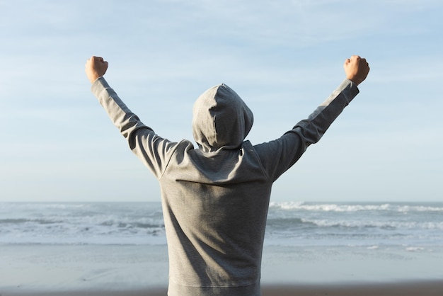 Achteraanzicht van een man met een hoodie die de overwinning of goede resultaten viert terwijl hij naar de zee kijkt