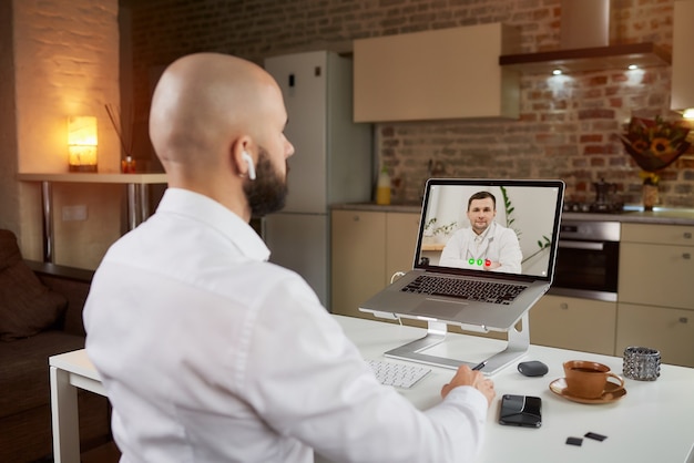 Achteraanzicht van een mannelijke werknemer in oortelefoons die naar een arts op een videoconferentie op een laptop luistert.