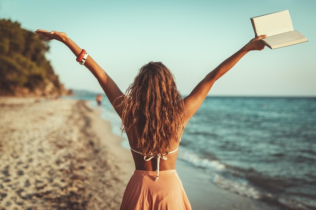 Achteraanzicht van een mooie jonge vrouw geniet op het strand met een boek in haar open armen.