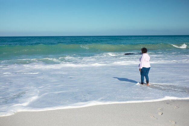 Achteraanzicht van een oudere Afro-Amerikaanse vrouw die op het strand staat met blauwe lucht en zee op de achtergrond en het water in loopt