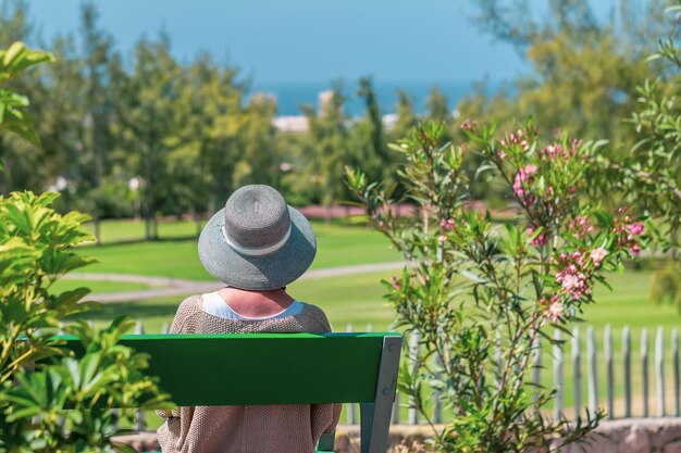 Achteraanzicht van een oudere blanke vrouw met hoed zittend op een bankje in het openbare park, kijkend naar de weide en de natuur Horizon over zee