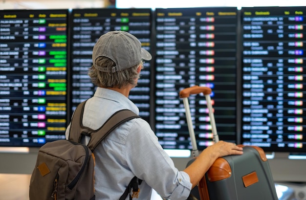 Achteraanzicht van een oudere vrouw op de luchthaven die naar het tijdschema kijkt om de vertrekpoort van haar vlucht te controleren Reiziger concept mensen met rugzak en koffers