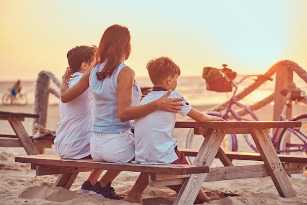 Achteraanzicht van een schattig gezin - moeder met haar zonen zittend op een bankje en kijkt naar de zonsondergang aan de zeekust.