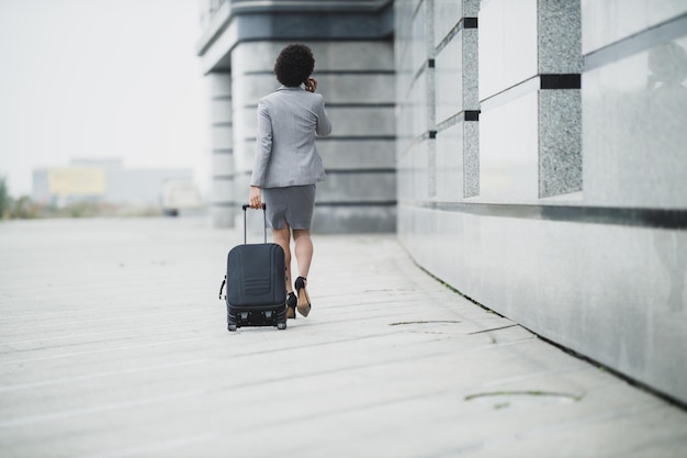 Achteraanzicht van een succesvolle zwarte zakenvrouw die met haar bagage loopt en een smartphone gebruikt buiten de luchthaven.