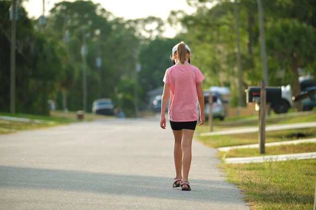 Achteraanzicht van een verdrietig jong kindmeisje dat alleen langs de groene straat loopt op een zonnige zomerdag