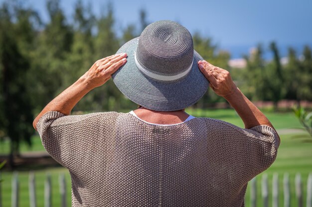 Achteraanzicht van een volwassen blanke vrouw die in een openbaar park staat en zijn hoed vasthoudt terwijl hij wegkijkt naar de horizon